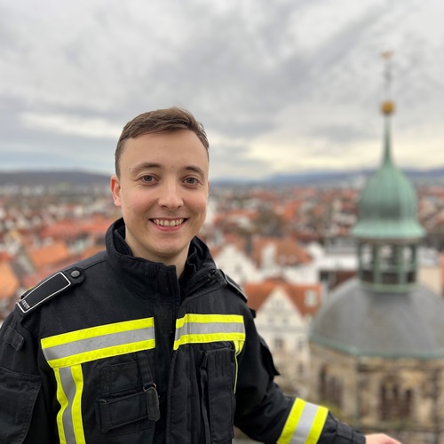 Junger Mann in Einsatzkleidung vor einer städtischen Kulisse.
