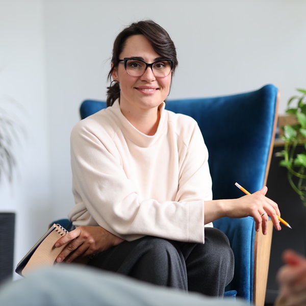 Porträt einer lächelnden Frau mit Brille und weißer Bluse, die auf einem Sofa sitzt.