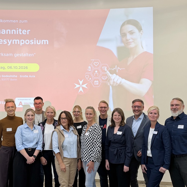 Gruppenfoto von Teilnehmenden des 2. Johanniter-Pflegesymposiums in der Johanniter-Klinik Godeshöhe in Bonn. Im Hintergrund ist eine Präsentation zum Thema Pflege zu sehen.