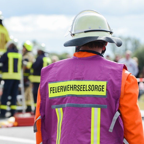 Einsatzkraft der Feuerwehr mit Helm und Warnweste, von hinten aufgenommen, an einer Einsatzstelle mit weiteren Einsatzkräften im Hintergrund.